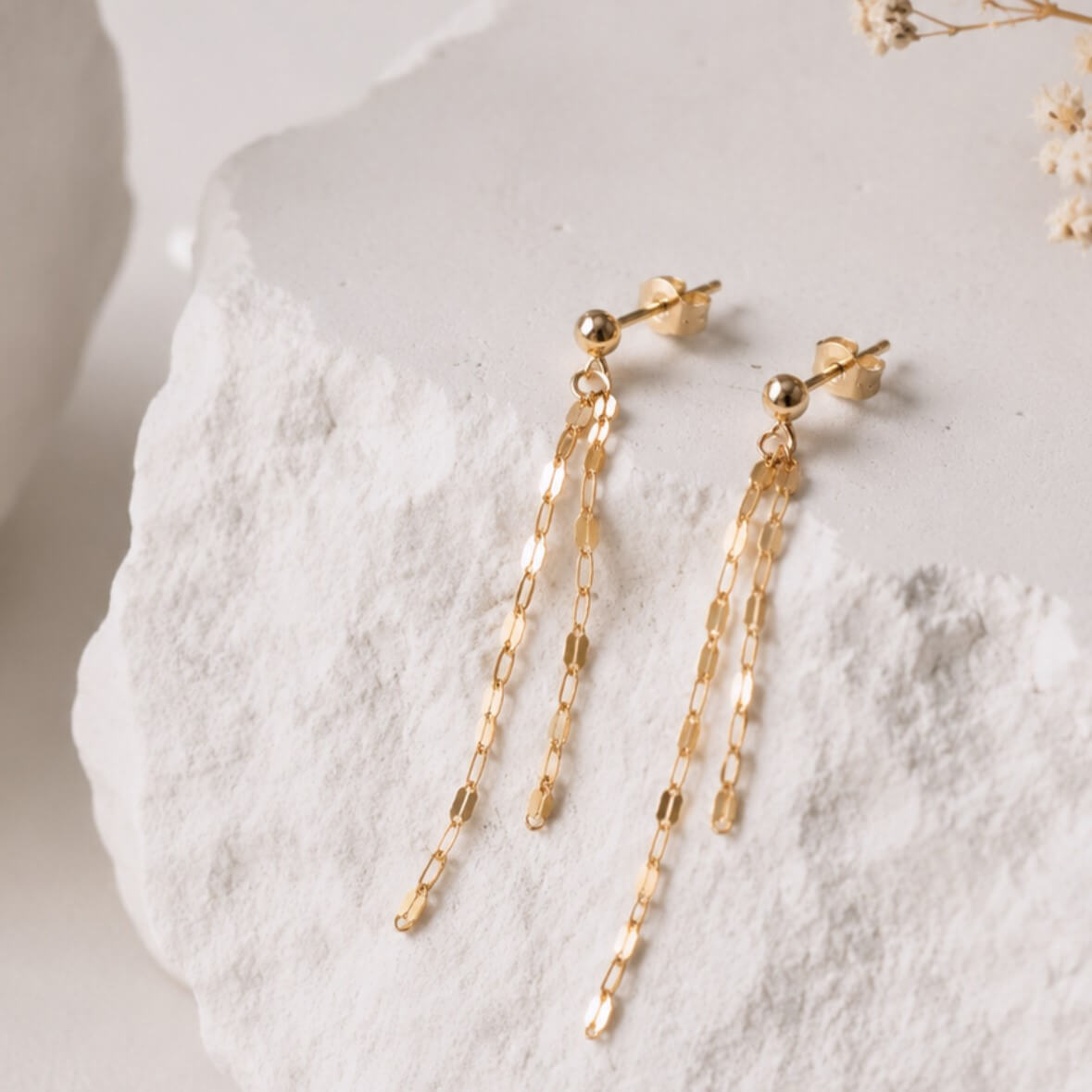 A pair of gold dangle oval disc chain earrings, displayed on a textured white stone surface.