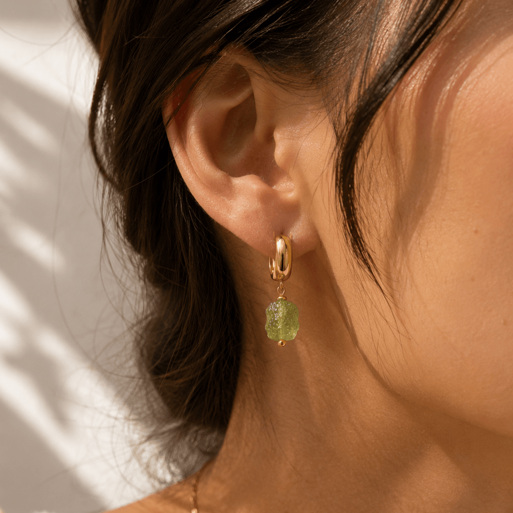 A close-up of a person's ear wearing a gold huggie hoop earring with a dangling textured light green gemstone.