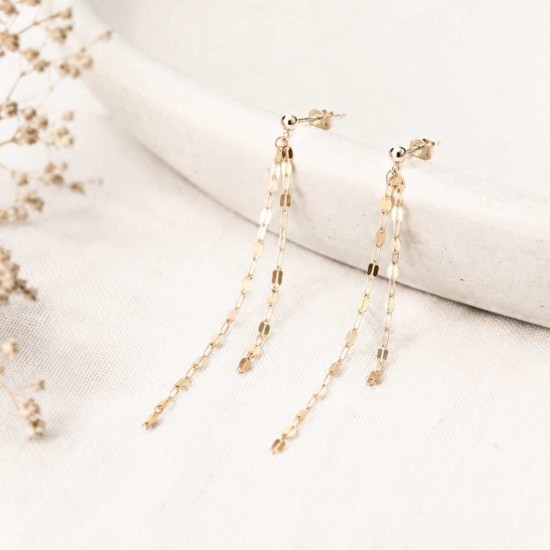 A pair of gold dangle earrings with multiple layered chain links and small disc charms, displayed on a textured white fabric next to a curved white stone prop with dried flowers.