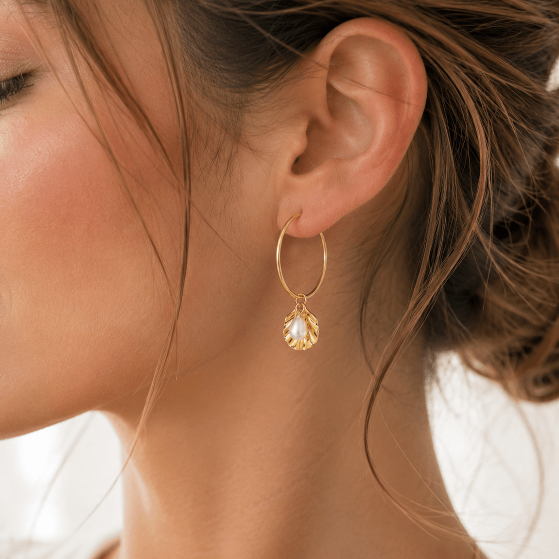 A close-up of a woman's ear wearing a delicate gold hoop earring with an oyster shell charm set with a rice pearl