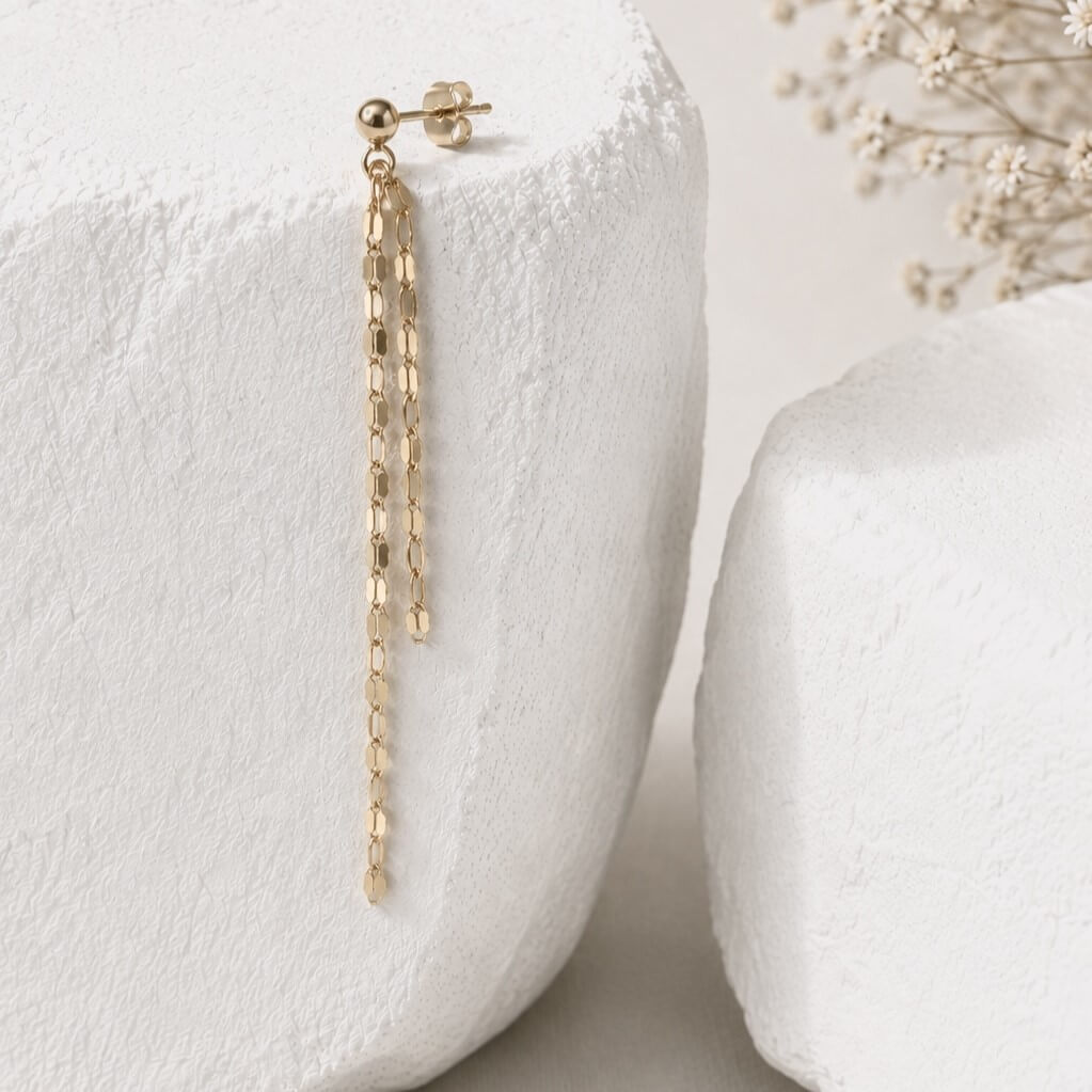 A delicate gold dangle earring with a short post and two long textured chain drops, displayed on a white textured stone block.