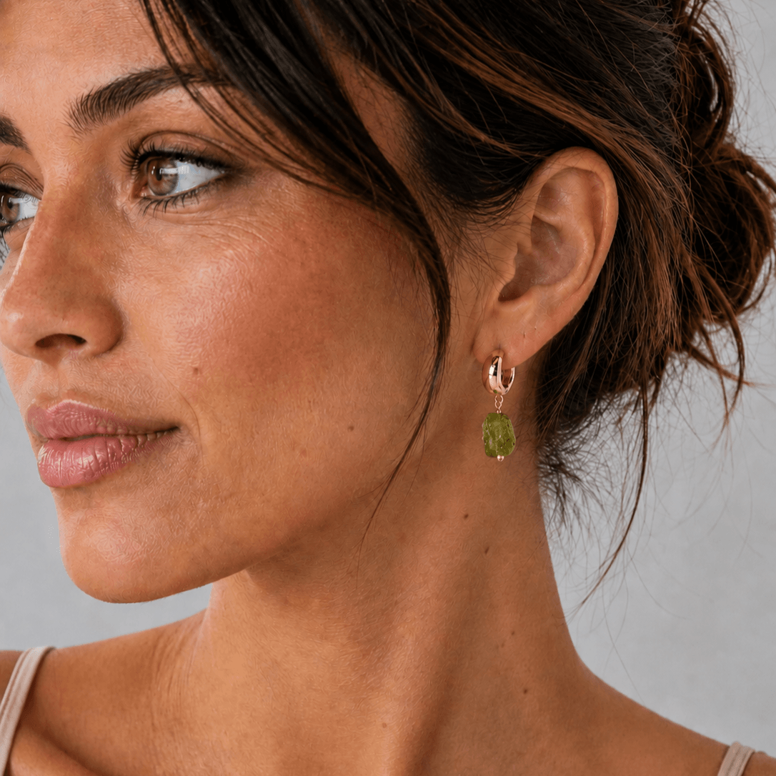 A close-up of a woman wearing a delicate gold huggie hoop earring with a small green stone dangle, shown from the side against a neutral gray background.