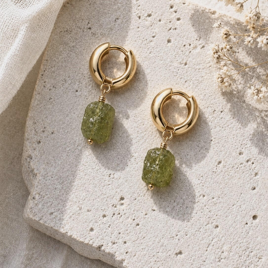 A pair of gold hoop drop earrings with natural raw green gemstone charm, displayed on a light beige stone surface with soft fabric and dried flowers.