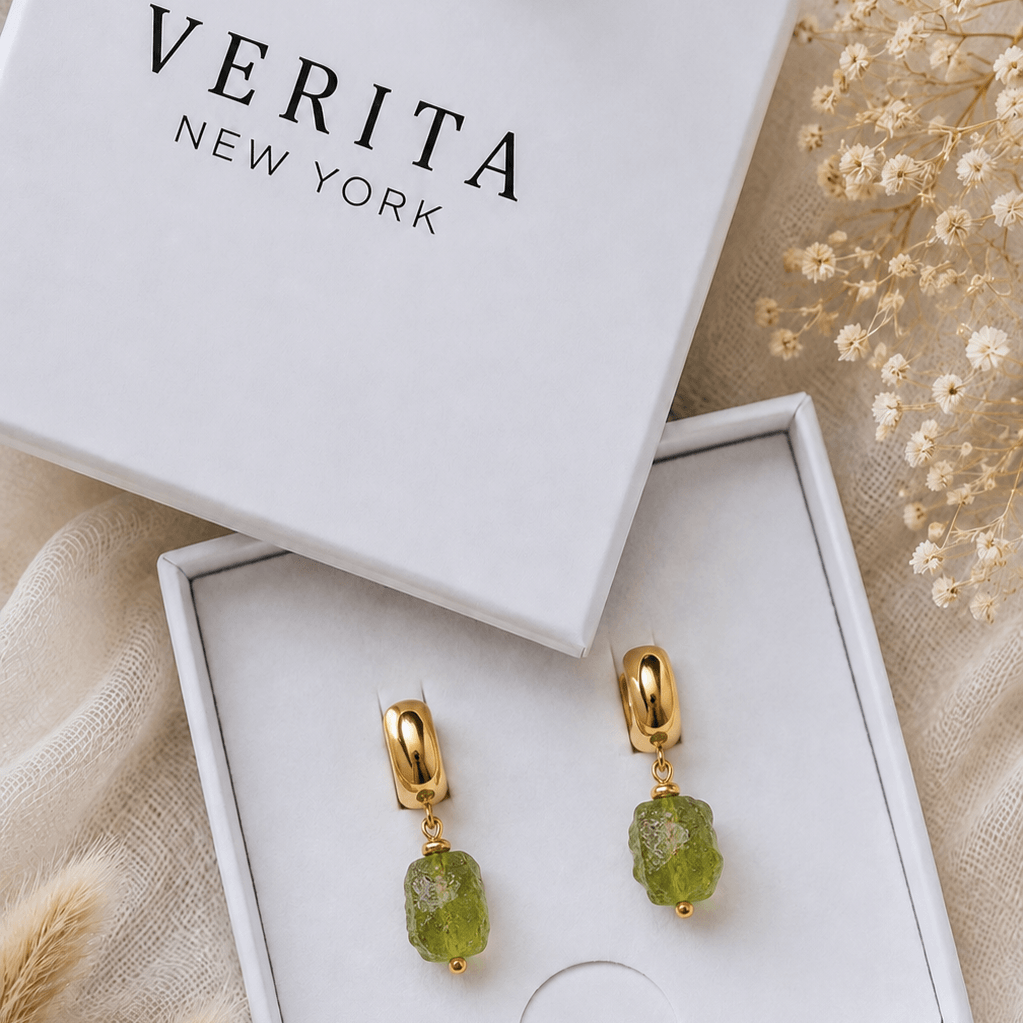 Verita New York gold drop earrings with green gemstone charms, displayed in an open white jewelry box next to the branded box lid on a beige fabric background with dried flowers.